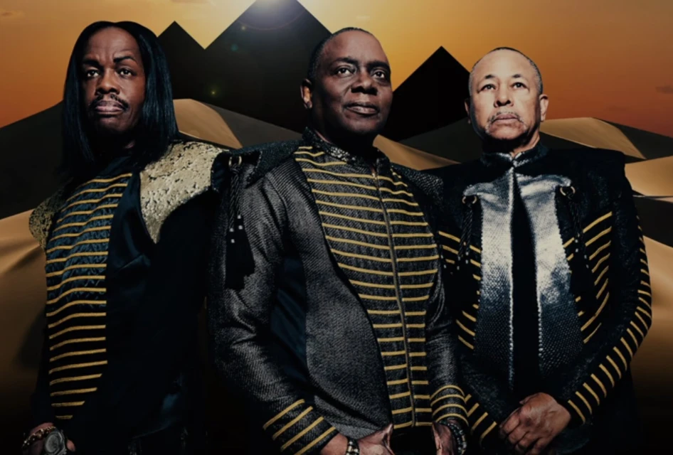 Three adult male musicians in black and gold striped jackets stand in front of a desert landscape with pyramids in the background.