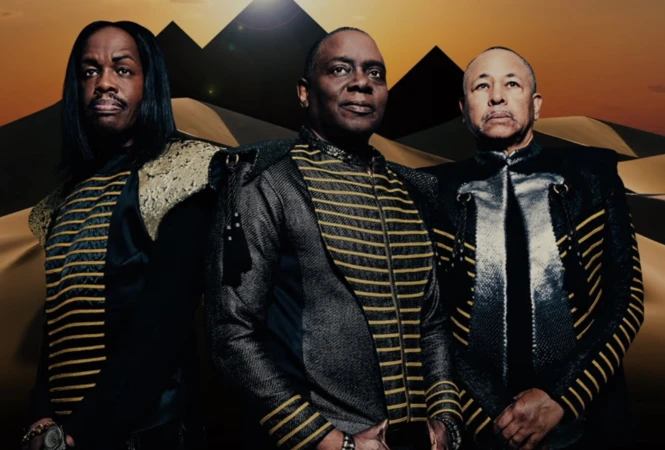 Three adult male musicians in black and gold striped jackets stand in front of a desert landscape with pyramids in the background.