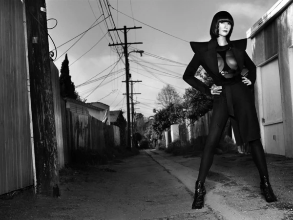 Black and white fashion shot of a woman in a coat and leggings posing in an alleyway with telephone poles and fences in the background.