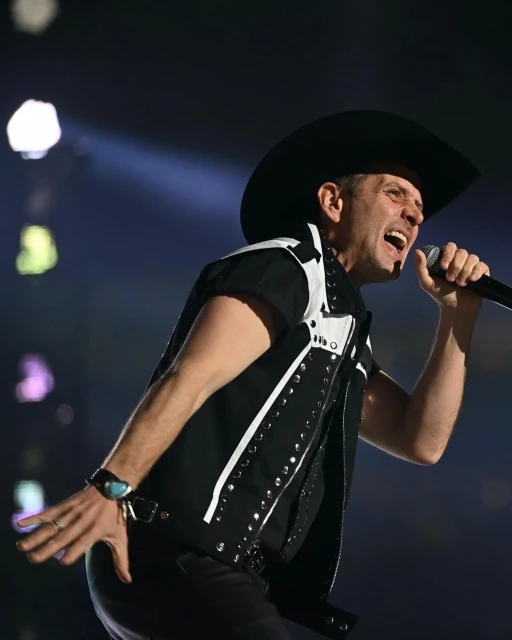 Adult male singer on stage wearing a black cowboy hat, singing into a microphone with a black vest and turquoise ring, spotlight.