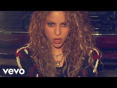 Close-up of Shakira with curly blonde hair, multiple necklaces, a black jacket, and a dark red truck in the background.