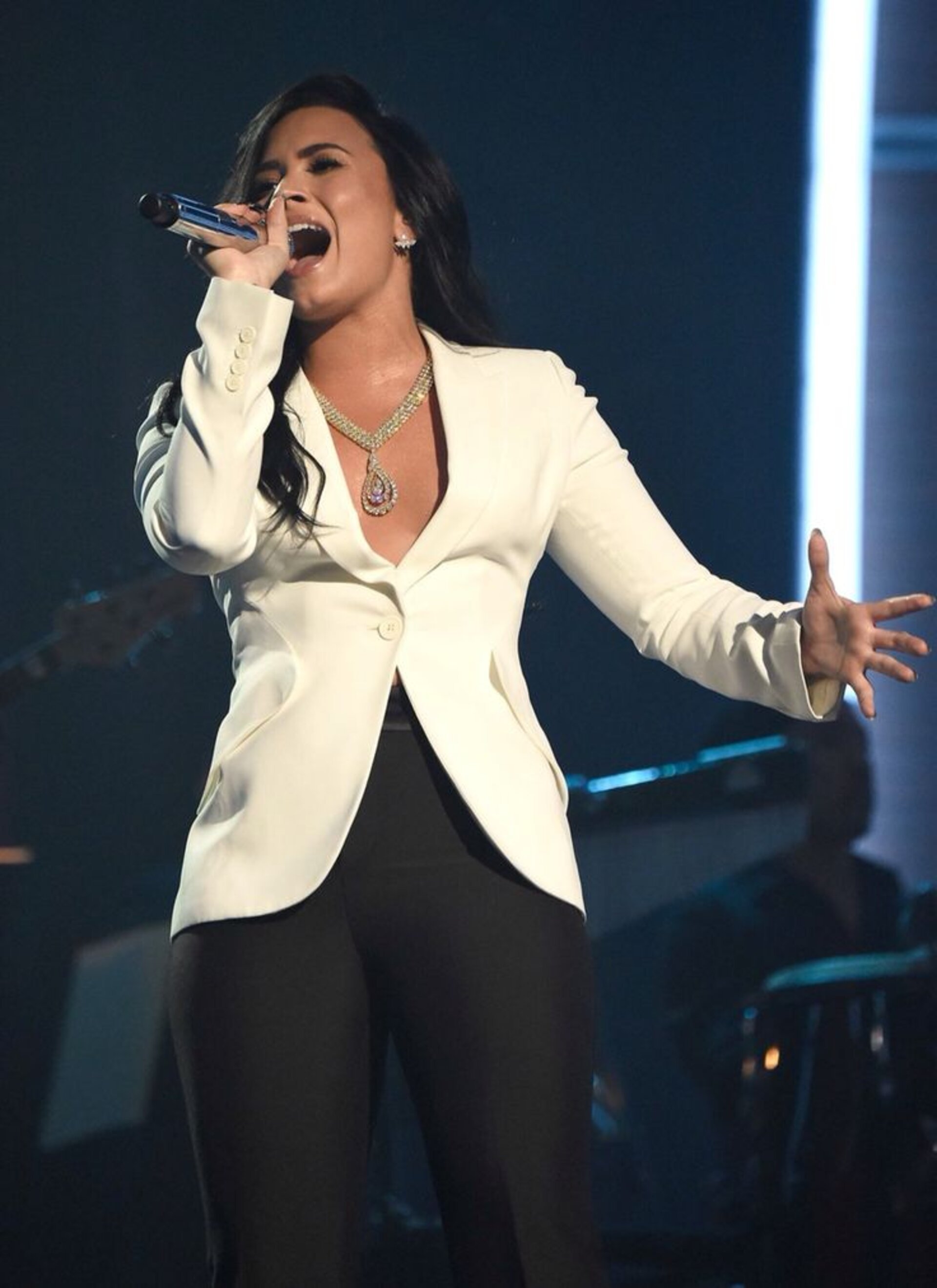 Demi Lovato performs on stage, singing into a microphone and wearing a white blazer and black pants, with band visible in the background.
