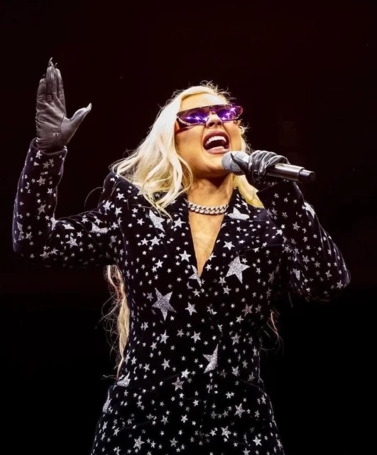 Christina Aguilera performs on stage, singing into a microphone while wearing sunglasses, a star-patterned blazer, and grey gloves.