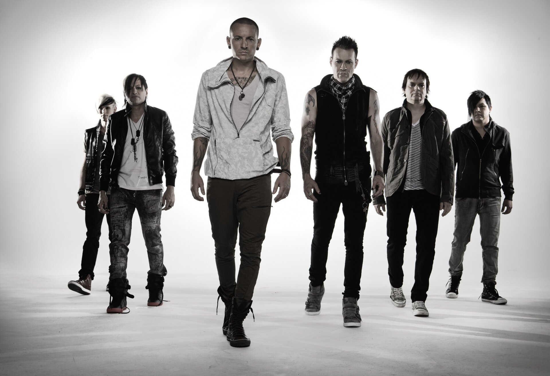 Group of six adult male musicians posing in a photo studio setting with white backdrop, wearing jackets, vests and casual wear, and dark shoes.