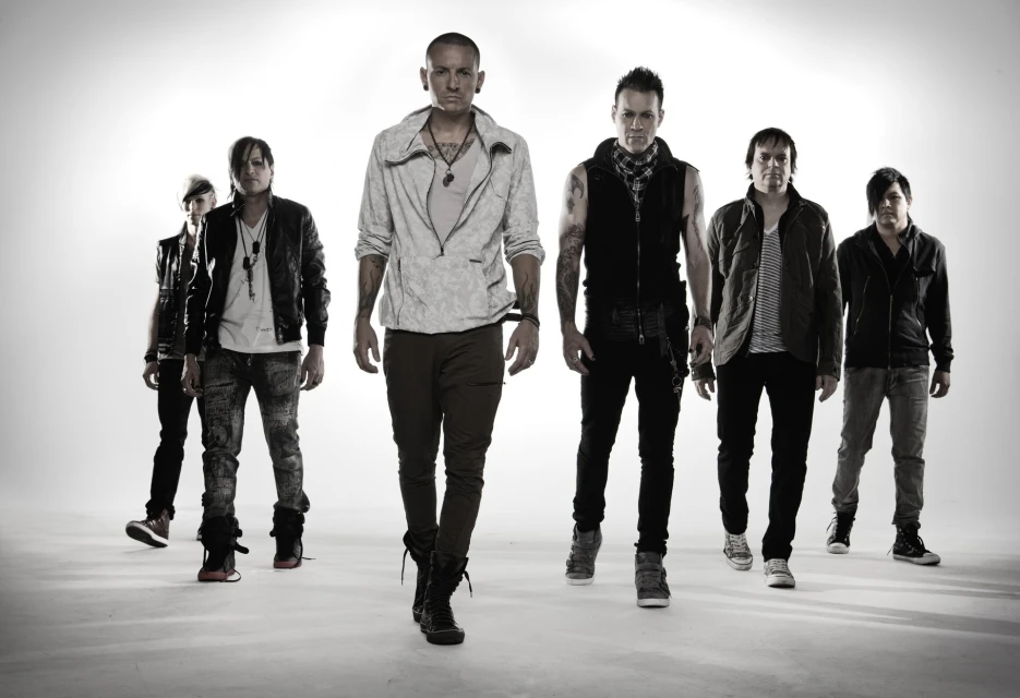 Group of six adult male musicians posing in a photo studio setting with white backdrop, wearing jackets, vests and casual wear, and dark shoes.