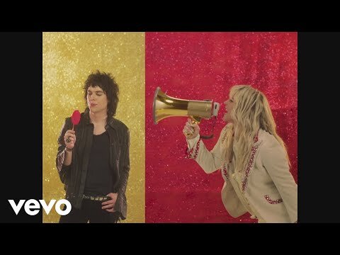 Two musicians, a male with a heart prop and a female with a megaphone, stand against a yellow and red glitter backdrop in a retro scene.