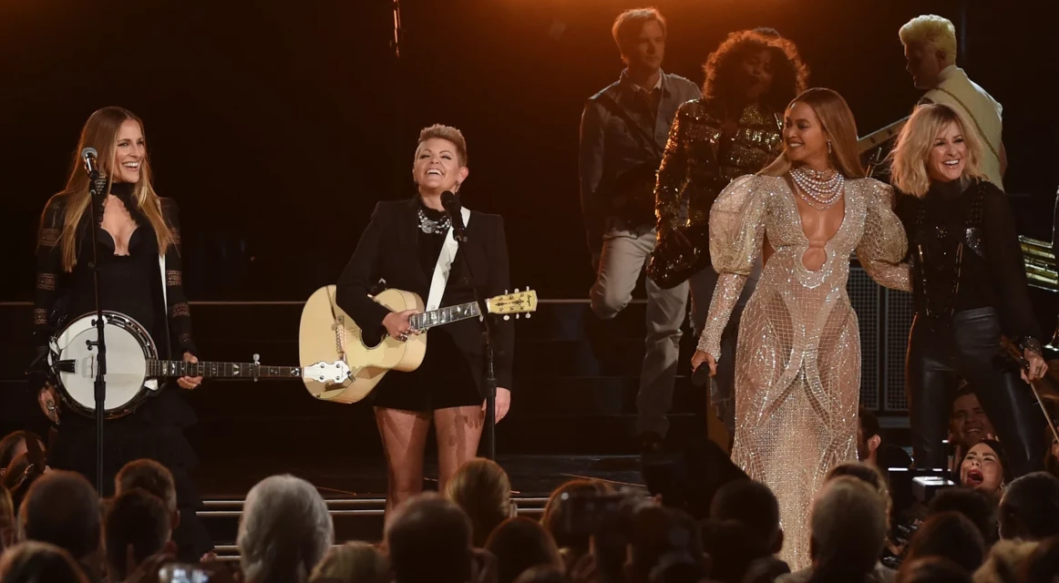 The Chicks and Beyoncé performing together on stage at an awards show, playing guitar, banjo, and violin in evening wear.