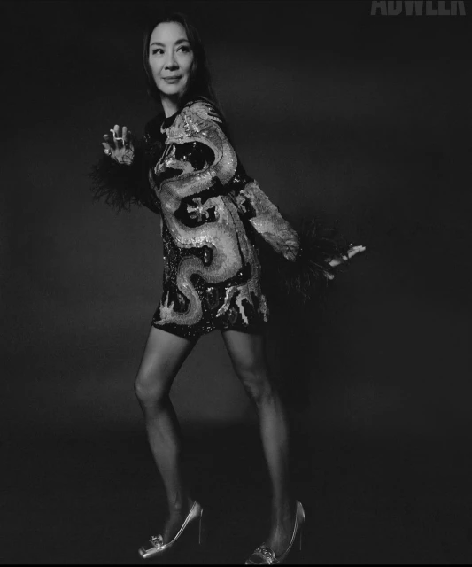 Michelle Yeoh poses in a sequined mini-dress with feathered sleeves, paired with sheer tights and heels, against a dark grey backdrop.