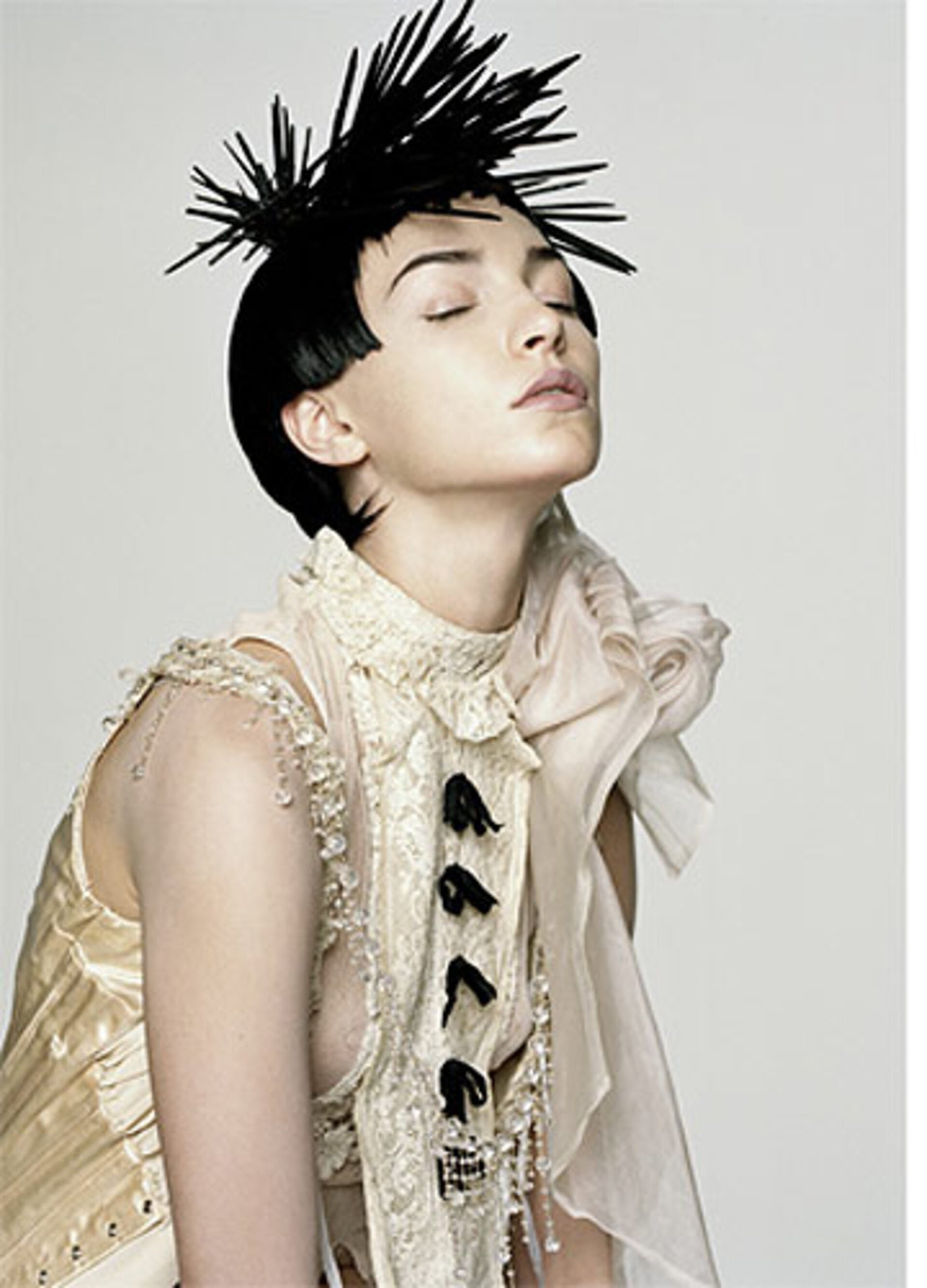 Woman with avant-garde black spike hair, eyes closed, wearing a textured cream-colored garment in a bright, soft, indoor studio setting.