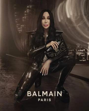 Cher poses in a shiny black suit with shoulder pads, holding a matching handbag.  The setting is a sepia-toned photo studio backdrop.