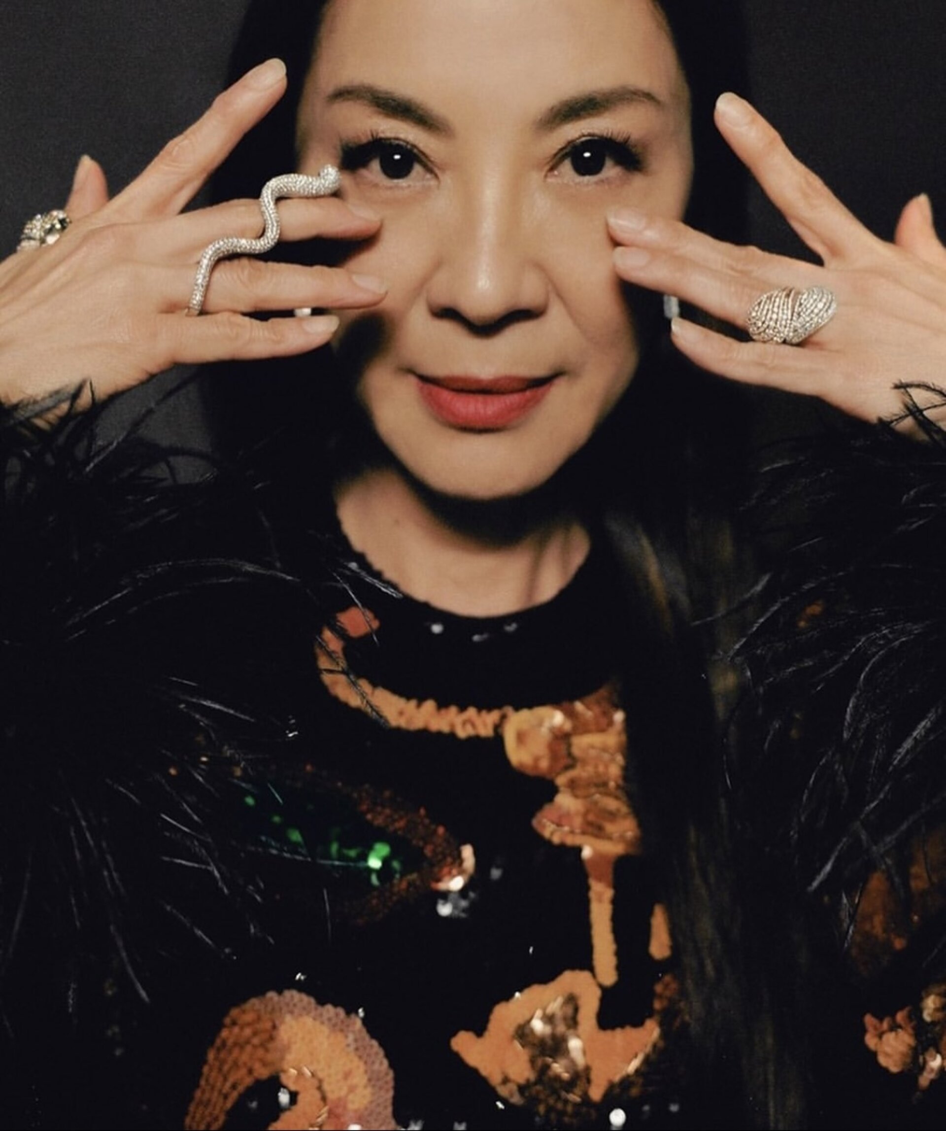 Close-up of Michelle Yeoh with hands near her face, showcasing unique rings, a sequined top, and feathery accents in a dark setting.