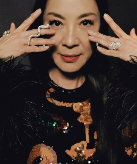 Close-up of Michelle Yeoh with hands near her face, showcasing unique rings, a sequined top, and feathery accents in a dark setting.