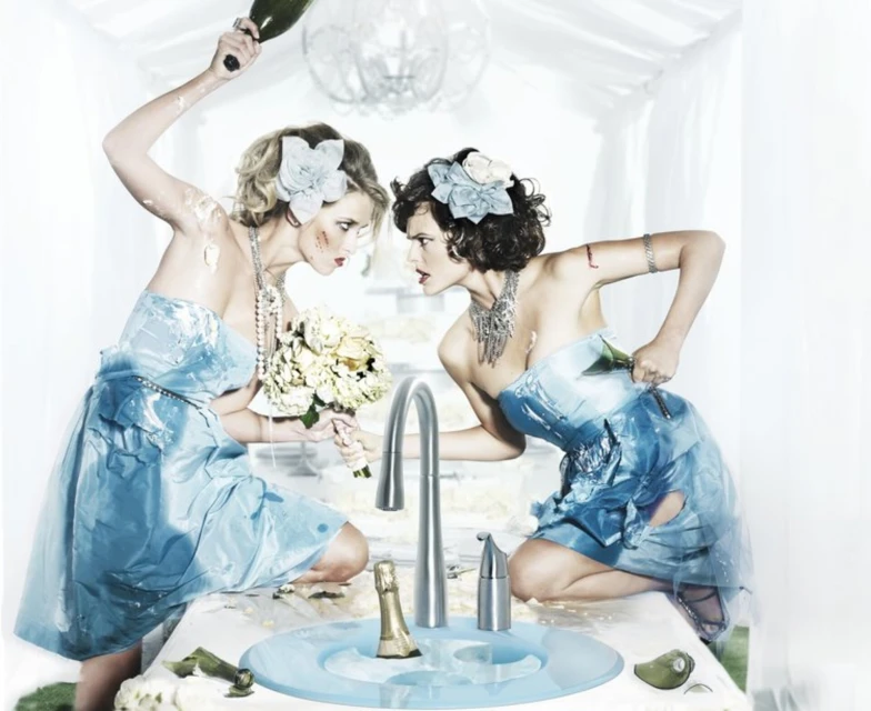 Two women in blue dresses, covered in cake, stand in front of a sink with a faucet, holding a champagne bottle and knife, in indoor setting.
