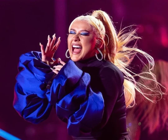 Singer Christina Aguilera performs on stage with an open mouth, raised hands, blue satin top, black turtleneck and blonde high ponytail.