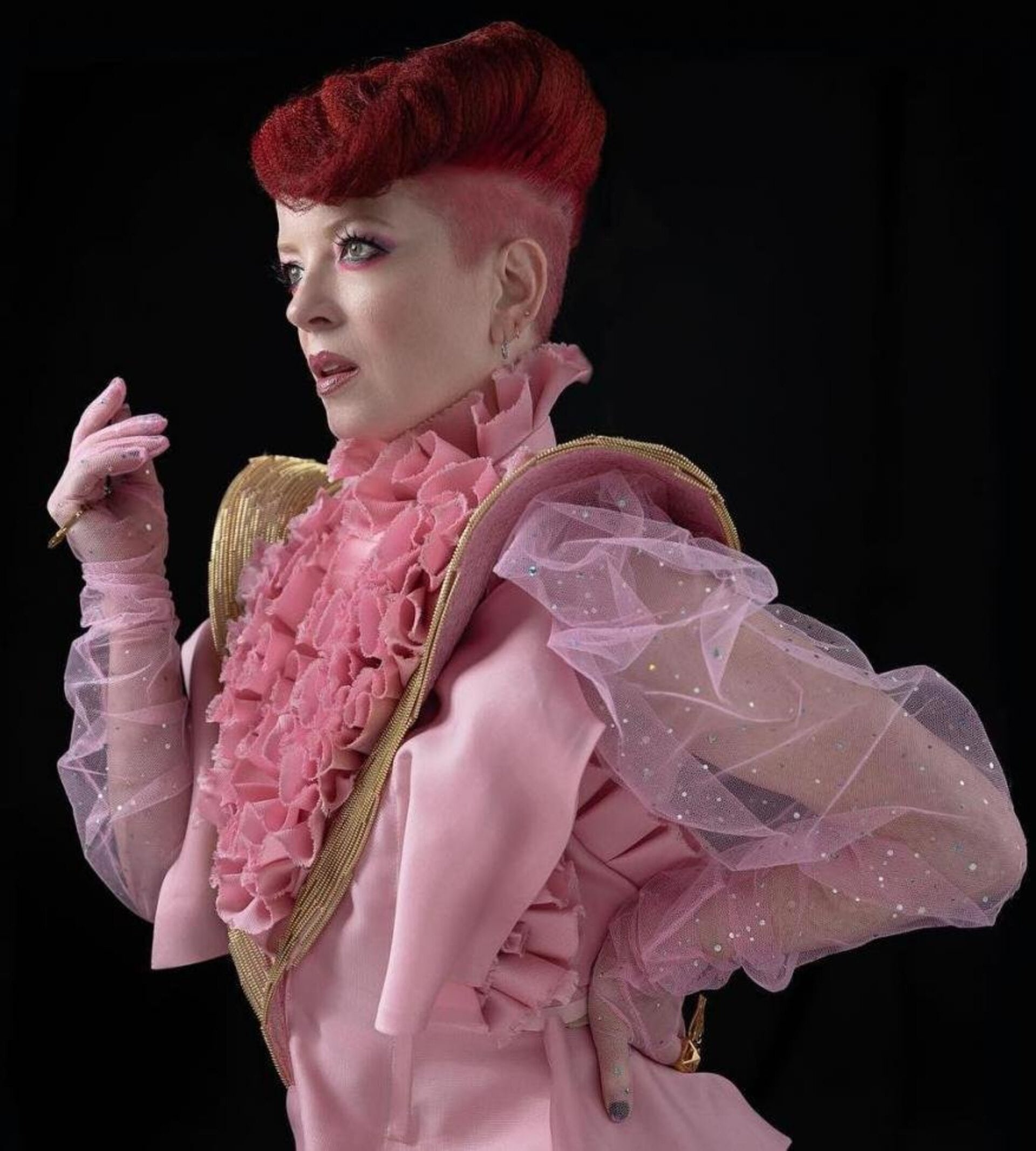 Elegant woman with red hair in pink dress, gold accents, and sheer sleeves poses in a studio against a black background. Beauty shot.