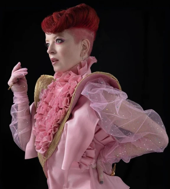 Elegant woman with red hair in pink dress, gold accents, and sheer sleeves poses in a studio against a black background. Beauty shot.