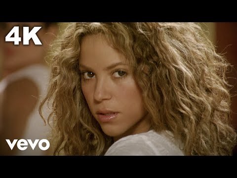 Shakira with voluminous curly blonde hair looking back over her shoulder in a white top.