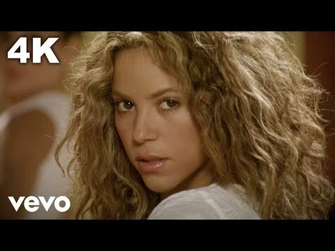 Shakira with voluminous curly blonde hair looking back over her shoulder in a white top.