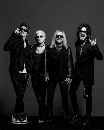 Four male rock musicians posing together in dark clothing and sunglasses against a plain grey studio backdrop.