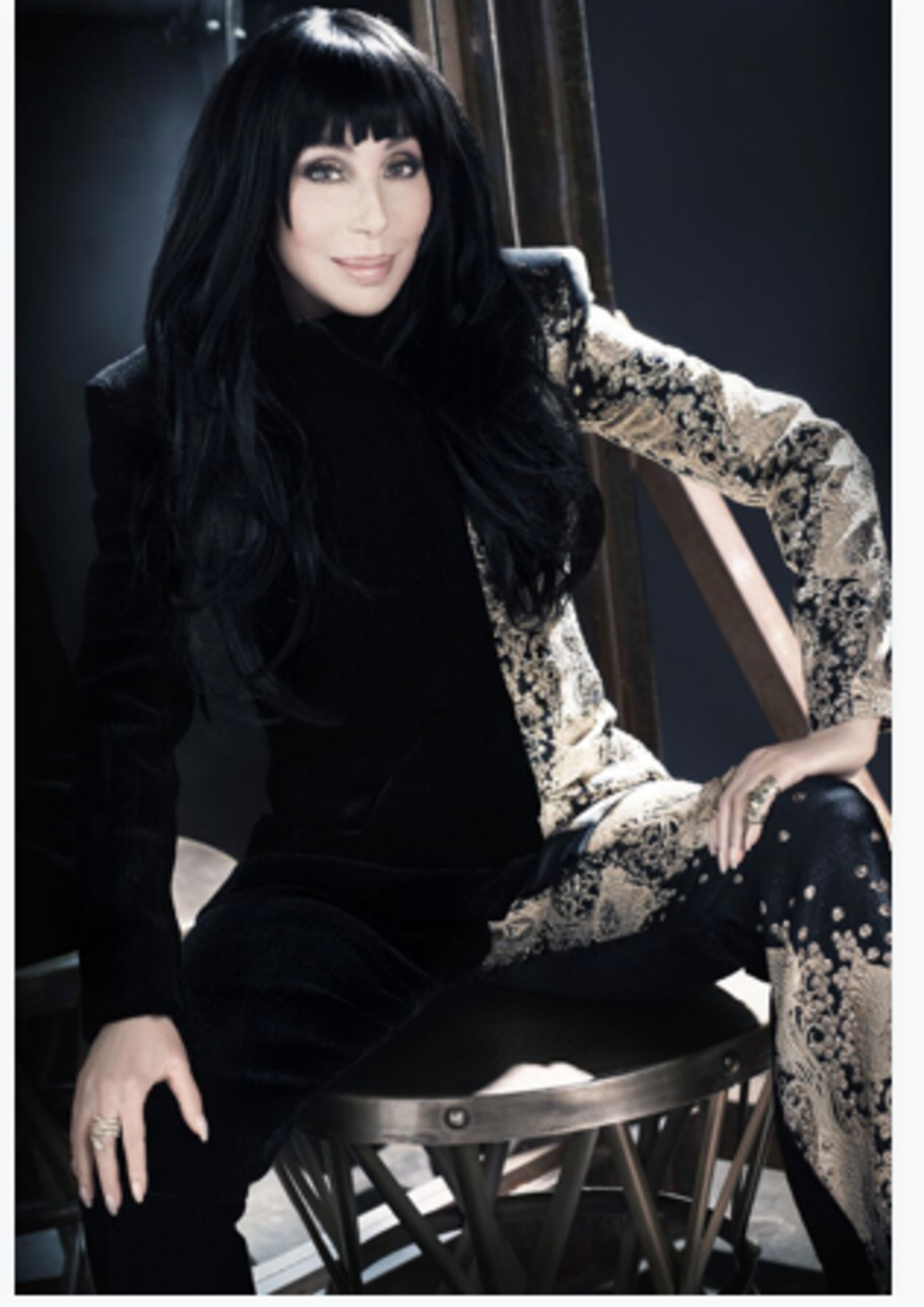 Portrait of Cher in a black and gold patterned suit, seated on a stool against a dark backdrop, showcasing a blend of elegance and style.