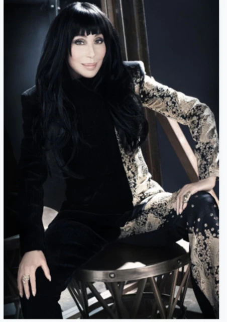 Portrait of Cher in a black and gold patterned suit, seated on a stool against a dark backdrop, showcasing a blend of elegance and style.