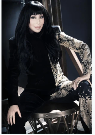 Portrait of Cher in a black and gold patterned suit, seated on a stool against a dark backdrop, showcasing a blend of elegance and style.