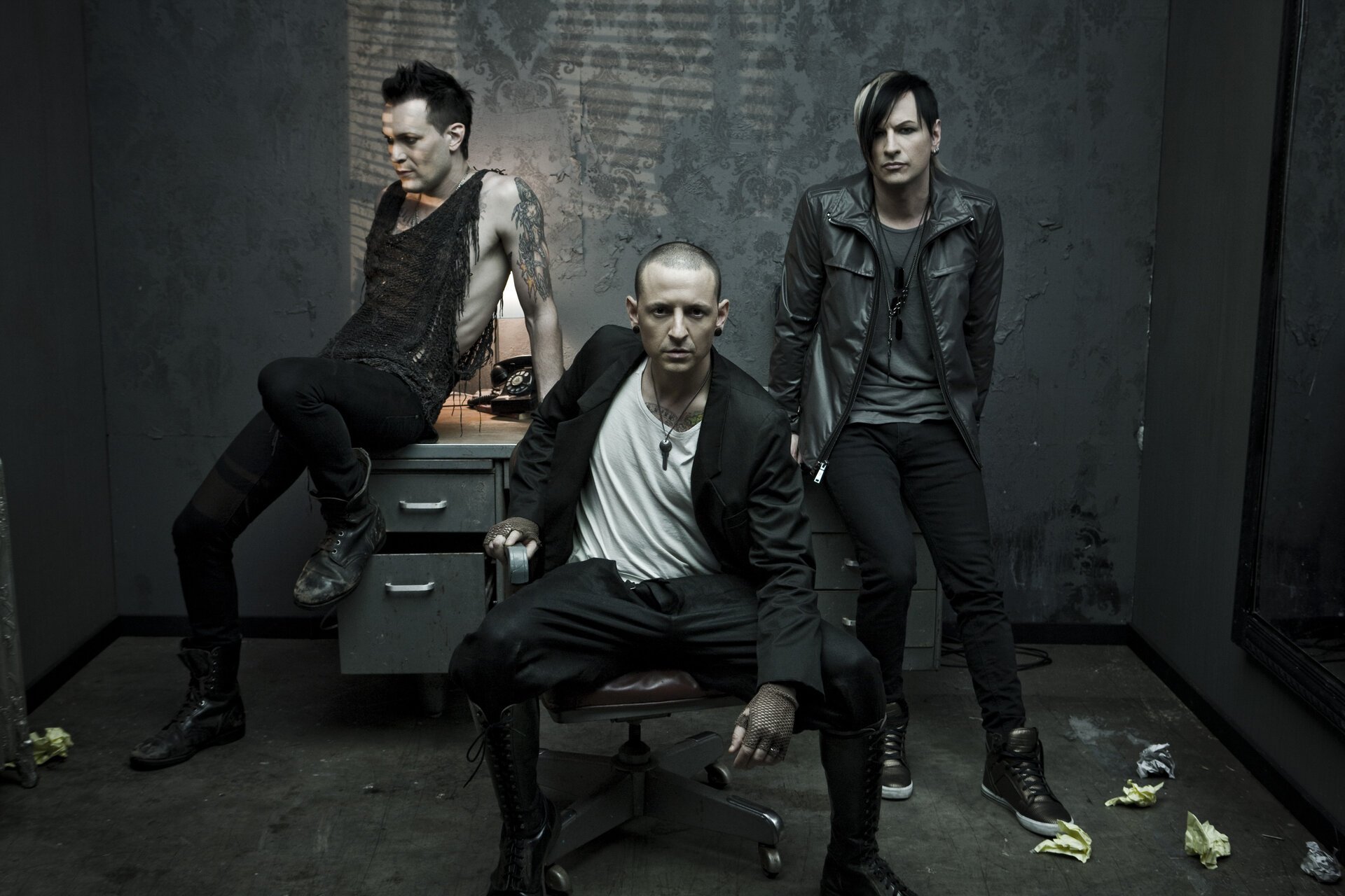 Three alternative rock band members pose in a dark, industrial-style room. One sits on a chair, one on a desk, one stands against a wall.