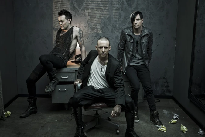 Three alternative rock band members pose in a dark, industrial-style room. One sits on a chair, one on a desk, one stands against a wall.