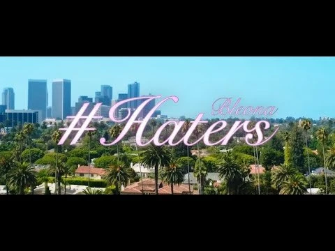 Cityscape with palm trees and high-rise buildings set against a clear blue sky and the hashtag 'Haters' in script font.