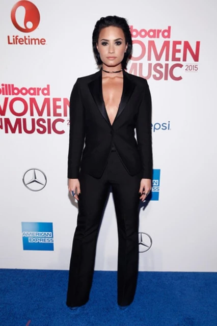 Demi Lovato attends Billboard Women in Music 2015 event, wearing black suit, against backdrop featuring event logos and sponsor signage.