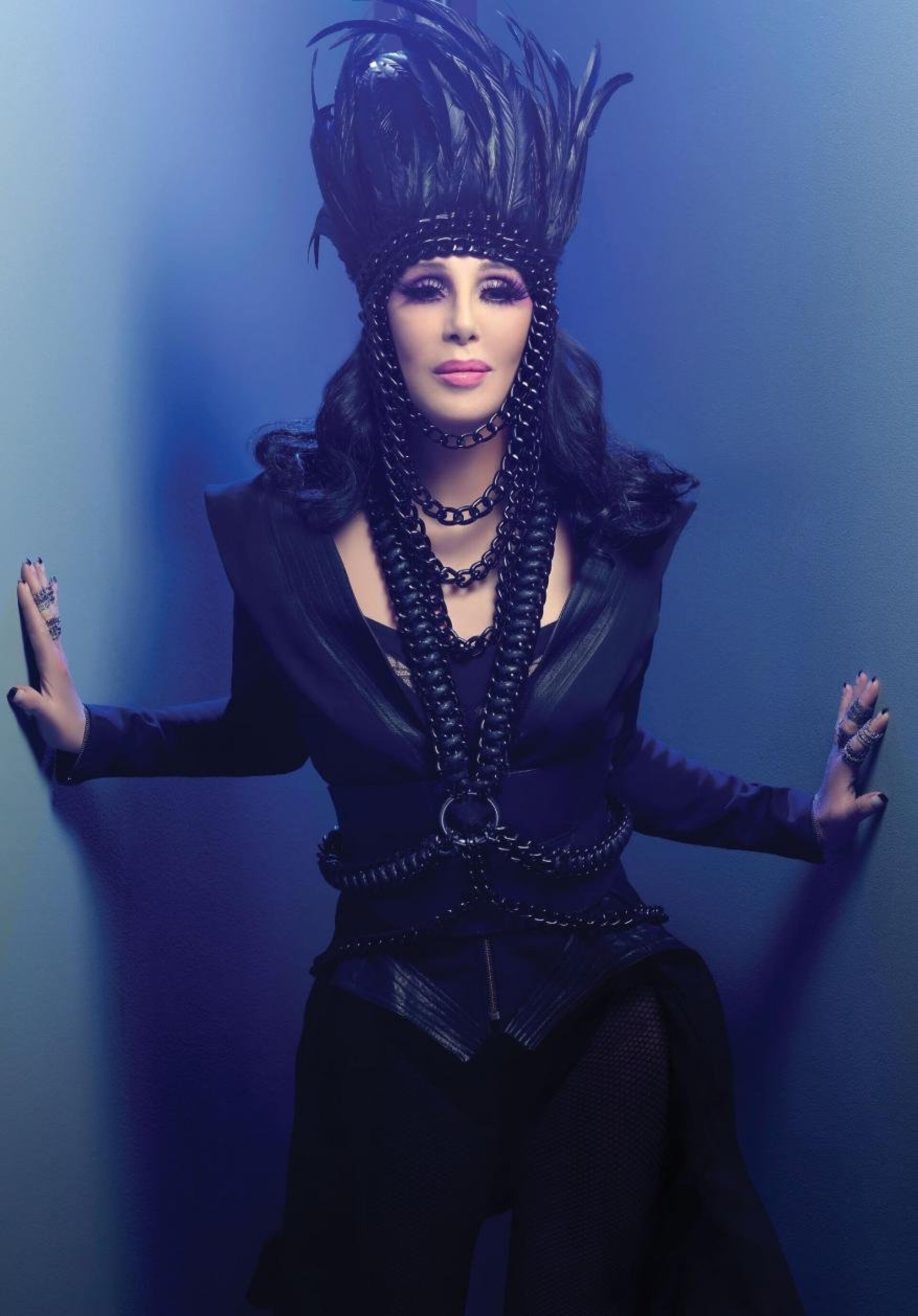 Portrait of a singer with black hair, wearing a black feather headdress, draped in chain necklaces, and posing against a blue wall.