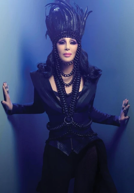 Portrait of a singer with black hair, wearing a black feather headdress, draped in chain necklaces, and posing against a blue wall.