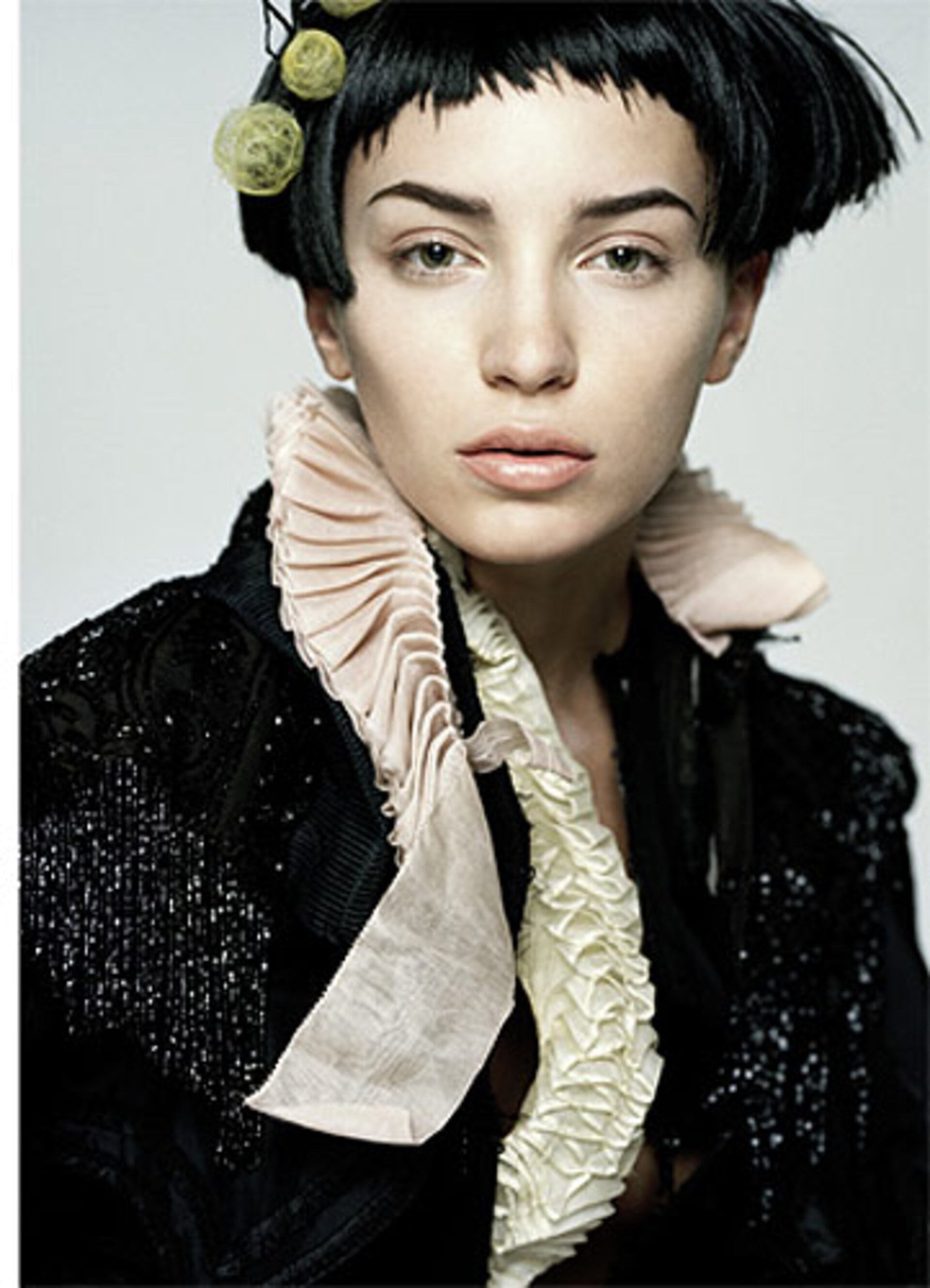 Portrait of a woman with short black hair wearing a black jacket with a ruffled collar and embellishments, shot against a plain background.
