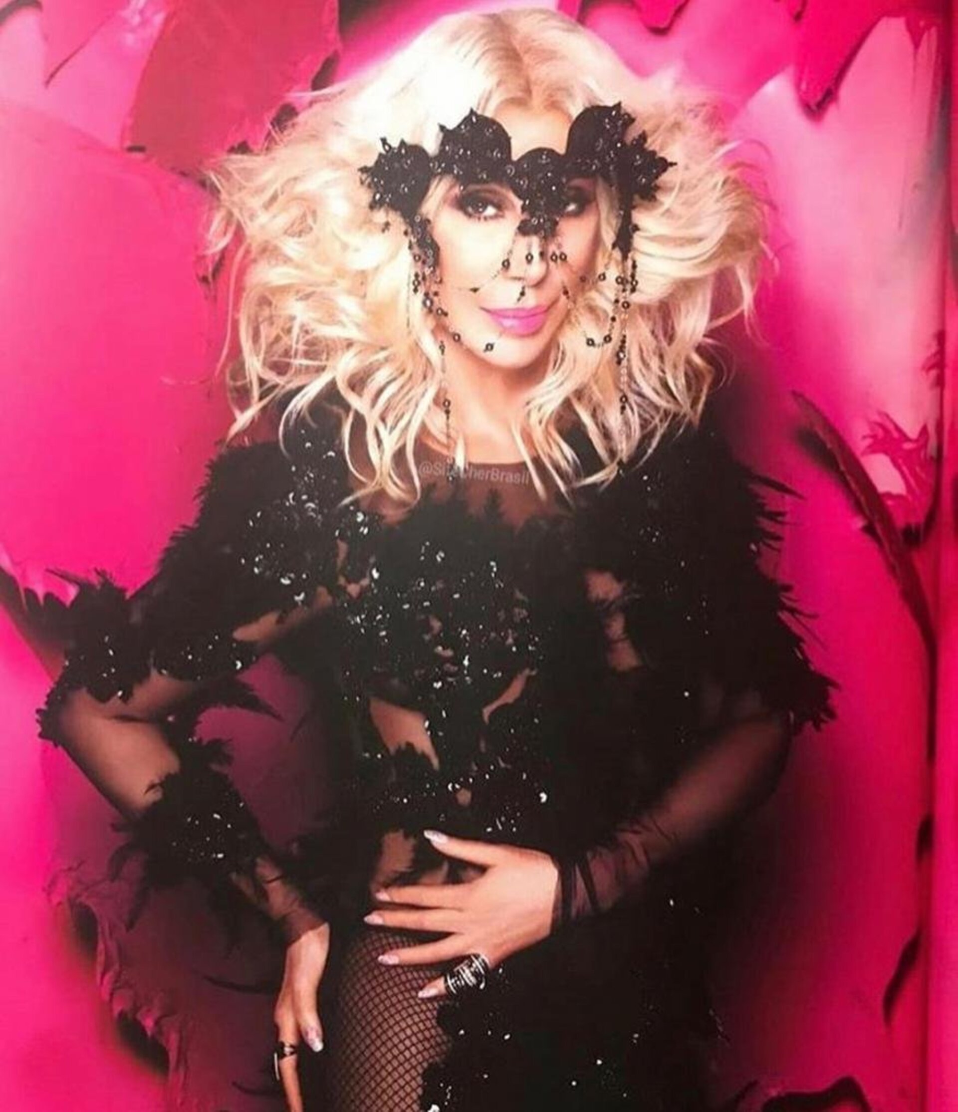Portrait of Cher wearing a black sequined dress, feather boa, and beaded headpiece, posing against a vibrant pink background. 