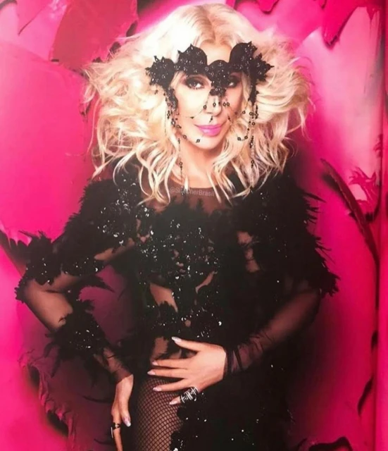 Portrait of Cher wearing a black sequined dress, feather boa, and beaded headpiece, posing against a vibrant pink background. 