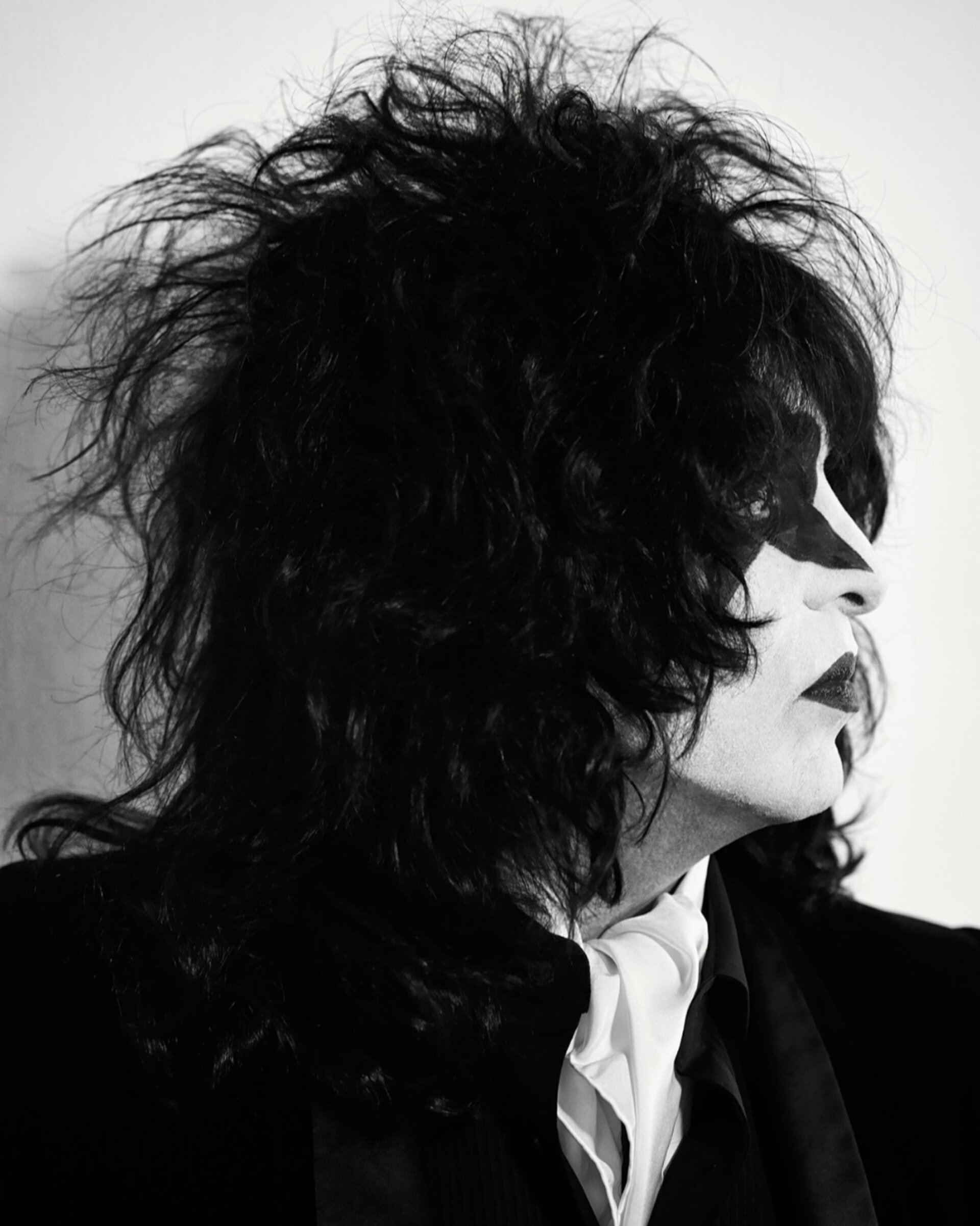 Black and white portrait of a person wearing face paint, with dark, tousled hair and a suit with a white cravat, against a plain background.