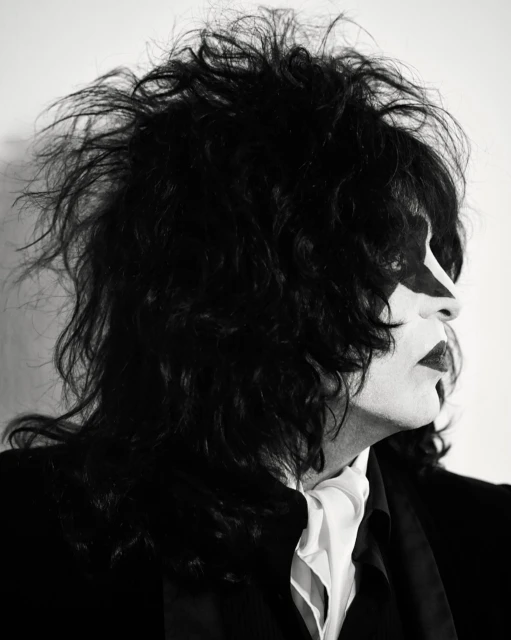 Black and white portrait of a person wearing face paint, with dark, tousled hair and a suit with a white cravat, against a plain background.