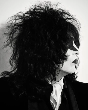 Black and white portrait of a person wearing face paint, with dark, tousled hair and a suit with a white cravat, against a plain background.