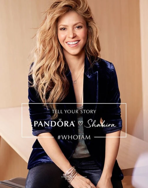 Close-up studio shot of Shakira wearing a navy velvet blazer with long wavy blonde hair and jewelry, promoting Pandora.
