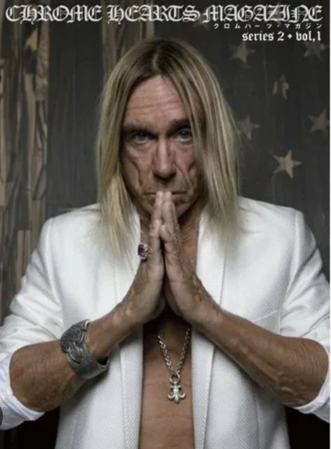 Close-up of Iggy Pop on a magazine cover with white blazer, silver jewelry, blonde hair, and a neutral expression in a photo studio.