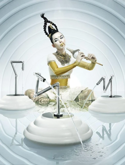 Asian woman with elaborate updo and makeup playing flute, seated in an artistic set with water features in an indoor environment.