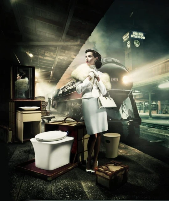 Elegant woman in vintage attire stands near luggage on a train platform next to a modern toilet, with a train and station backdrop.