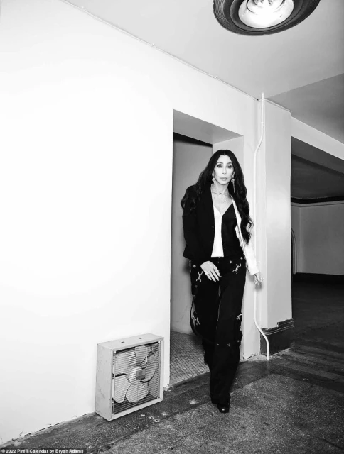 Cher in black and white portrait. She wears a blazer, pants with white designs, and is inside a building with white walls.