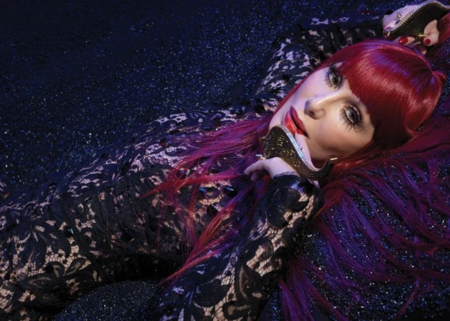 Portrait of a woman with red hair wearing black lace dress, reclining on a textured black surface in studio lighting for a fashion shoot.