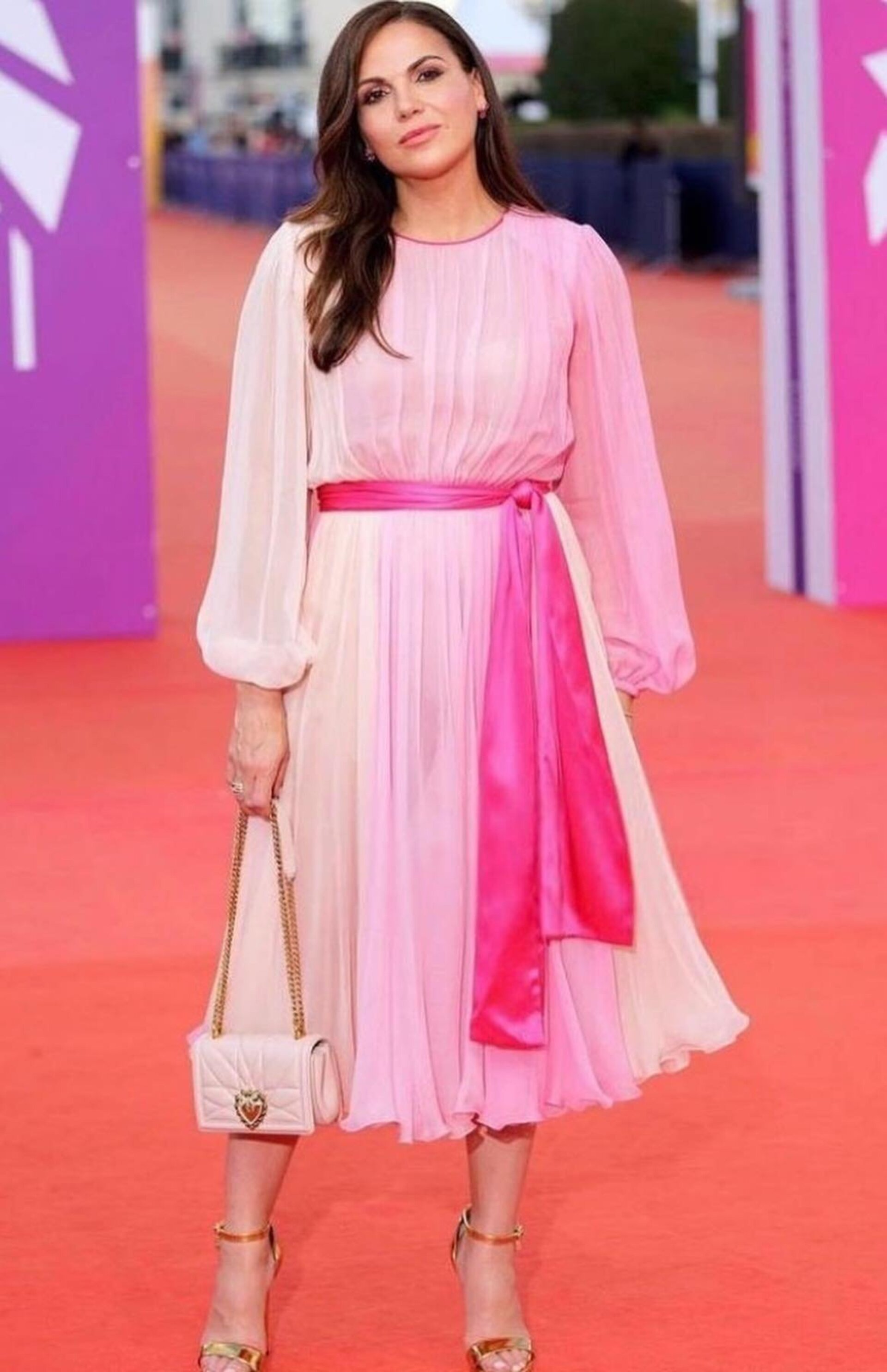 Elegant woman in a pastel pink dress with a matching purse and gold heels, standing on a red carpet in an outdoor setting.