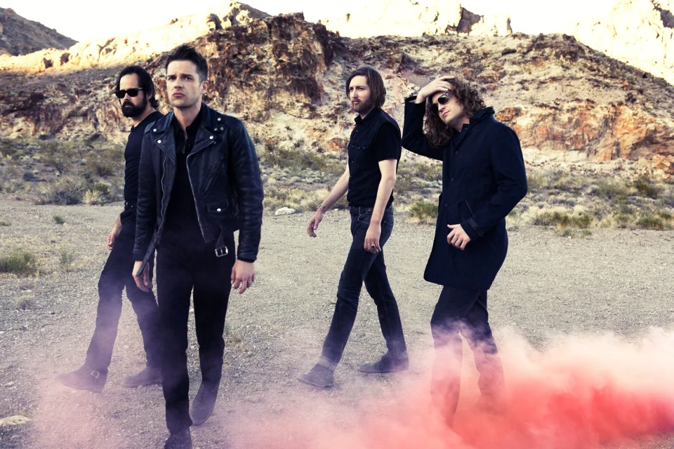 Four men in black clothing walk across a desert landscape filled with red smoke, backed by rocky mountains and a clear sky.