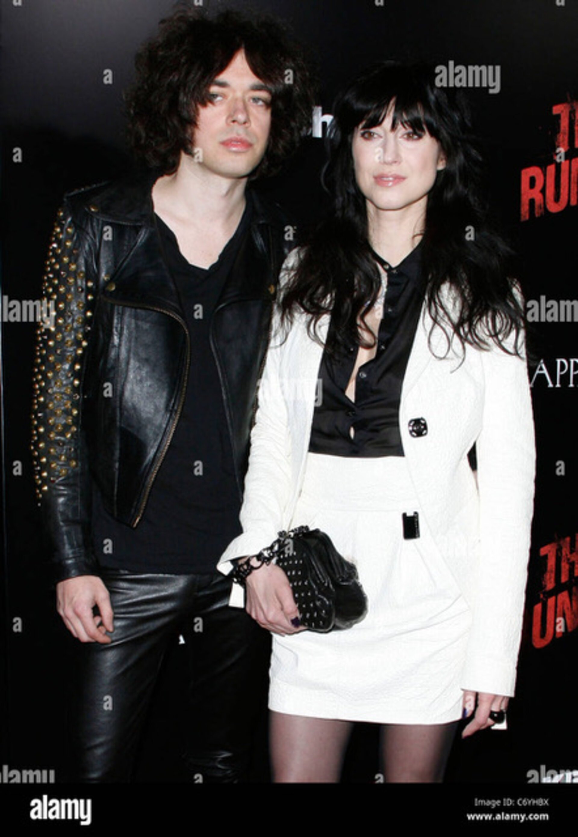 Stylish photo of a man in a studded leather jacket with a woman in a white suit at a premiere, both posing against a dark backdrop.