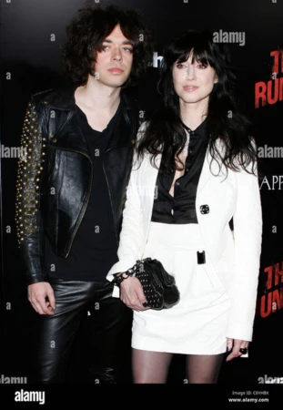 Stylish photo of a man in a studded leather jacket with a woman in a white suit at a premiere, both posing against a dark backdrop.