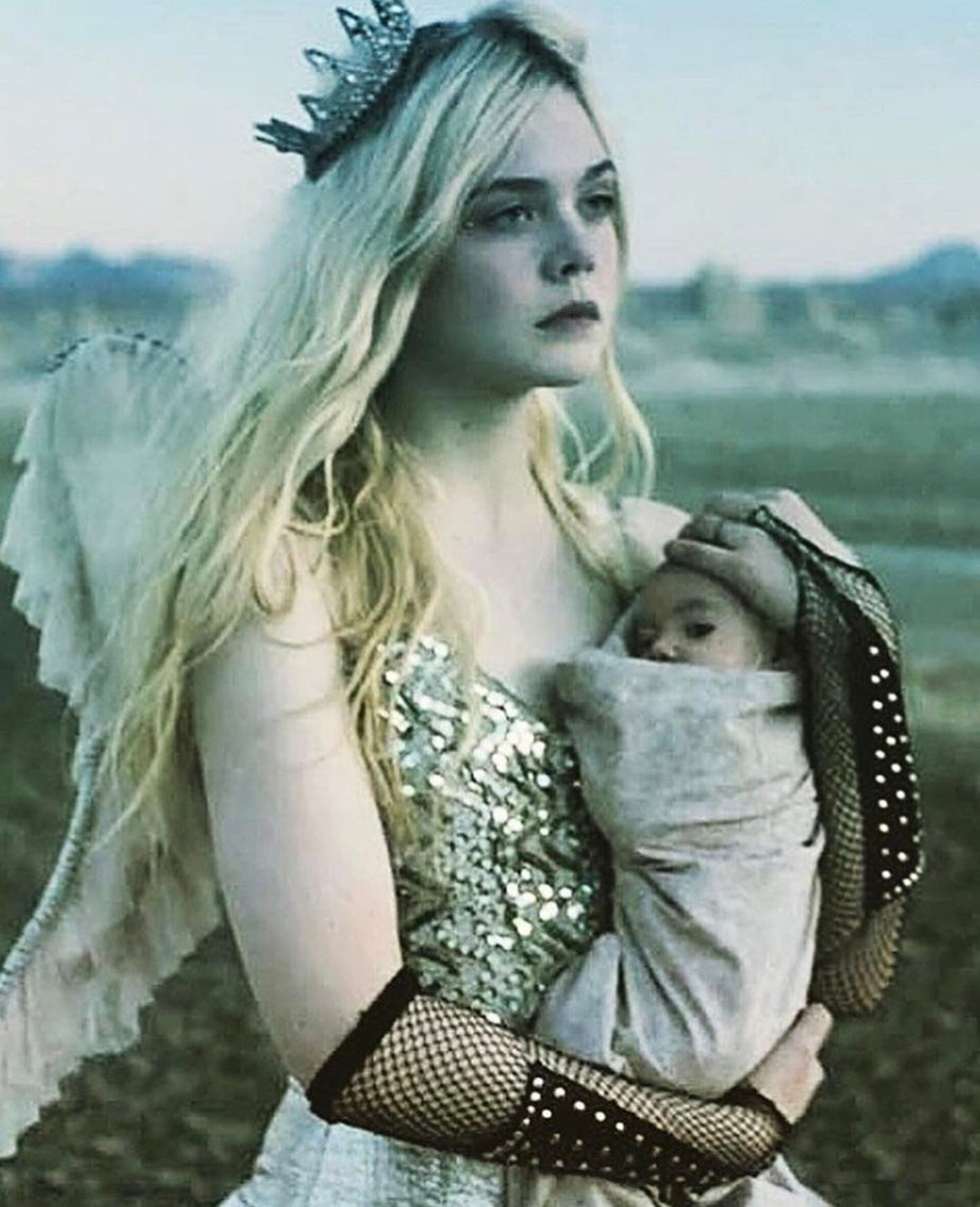 Elle Fanning, wearing a crown and angel wings, holds a baby wrapped in fabric. The background shows a blurred landscape and sky.