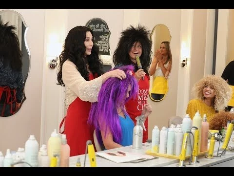 Cher tending to a woman with purple hair, while a woman in an afro wig holds a microphone, in a brightly lit beauty salon setting.